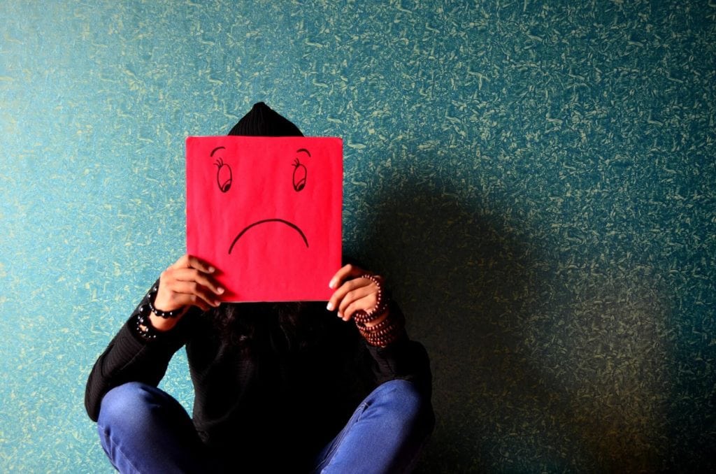 A photograph of a person sitting down, holding a red paper with a sad face drawn on it in front of their actual face.